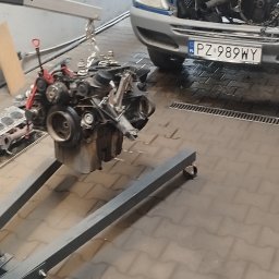 CarService- Dominik Muszkieta - Wyjęty silnik zawieszony na łańcuchu na tle niebieskiego samochodu z otwartą maską w warsztacie, widoczne narzędzia na ścianie.