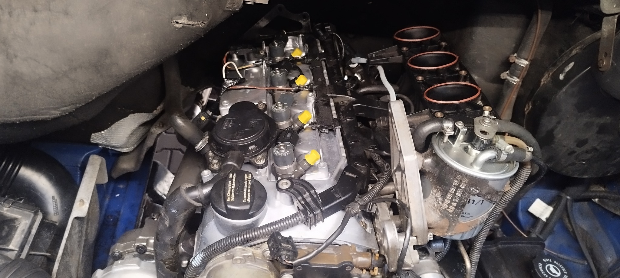 Widok z góry na silnik spalinowy z widocznymi wtryskiwaczami, przewodami, filtrem paliwa i pokrywą wlewu oleju, umieszczony w niebieskiej komorze silnikowej, z elementami osłon.