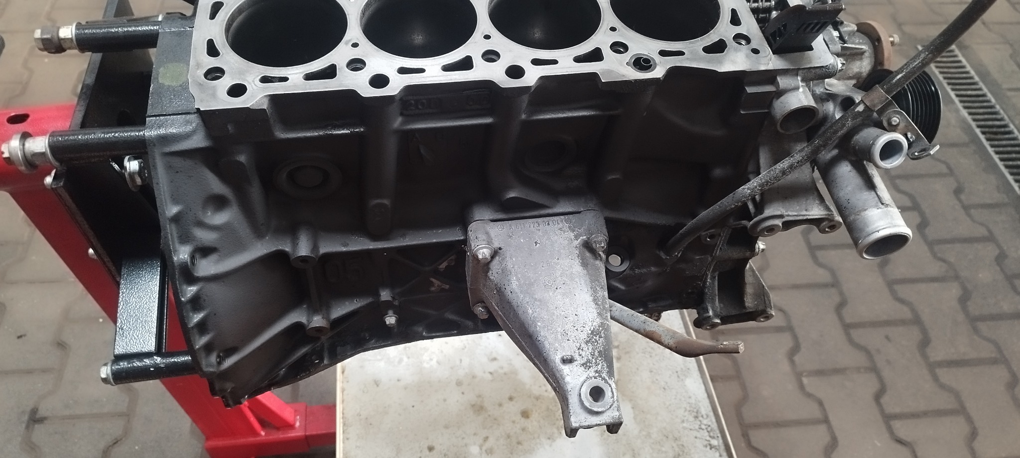 Czarny blok silnika samochodowego z widocznymi otworami cylindrów, zamontowany na czerwono-czarnym stojaku warsztatowym, oświetlony światłem dziennym.