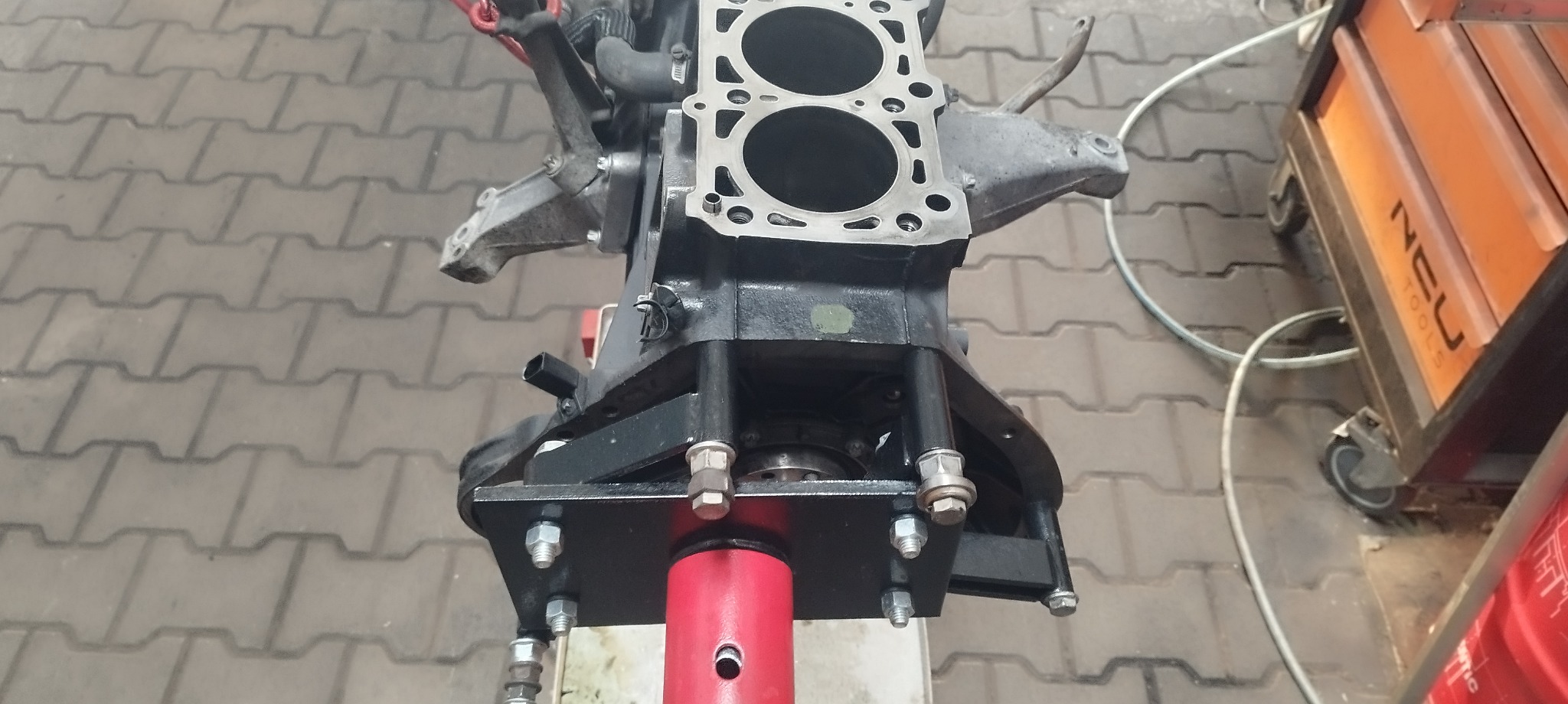Zdemontowany blok silnika zamontowany na czerwonym stojaku warsztatowym, na tle kostki brukowej i wózka narzędziowego NEO Tools.