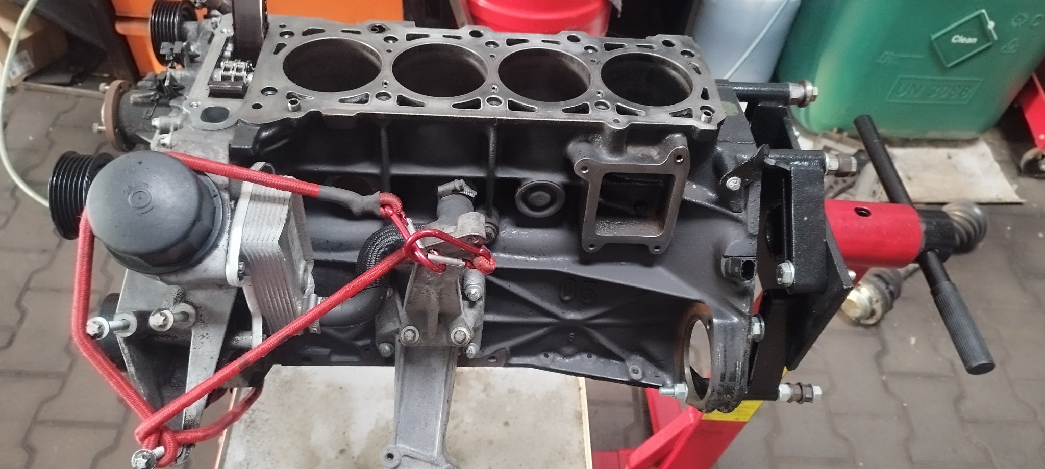 Widok z góry na zdemontowany, czarny blok silnika na czerwonym stojaku warsztatowym, zabezpieczony czerwoną liną, z widocznymi otworami cylindrów i elementami osprzętu.