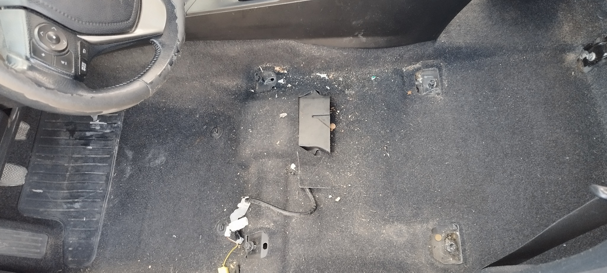 Wnętrze samochodu zdemontowane, widoczny brudny dywanik podłogowy z elementami mocującymi i przewodami, fragment kierownicy po lewej stronie.