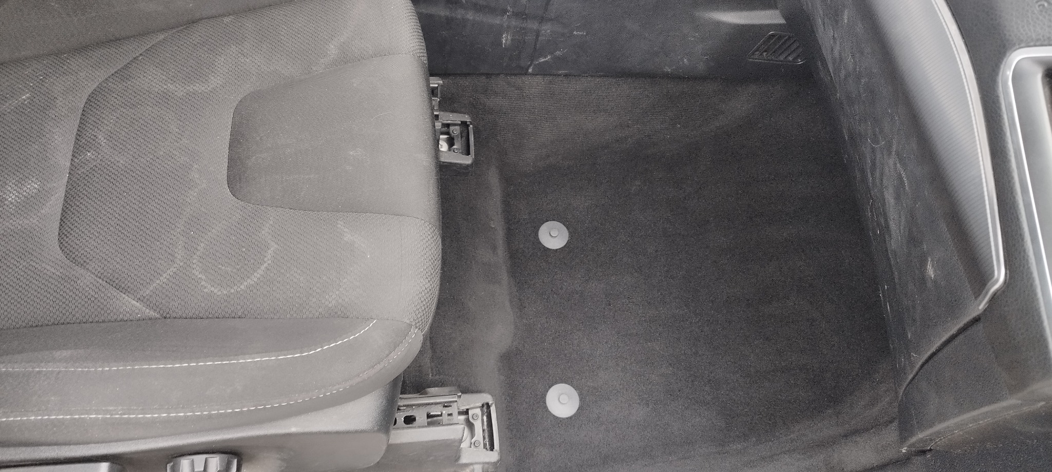 Wnętrze samochodu: czarne tekstylne siedzenie z widocznym mechanizmem regulacji, czarna podłoga z dwoma okrągłymi zaczepami dywaników, widoczne zabrudzenia i ślady użytkowania.