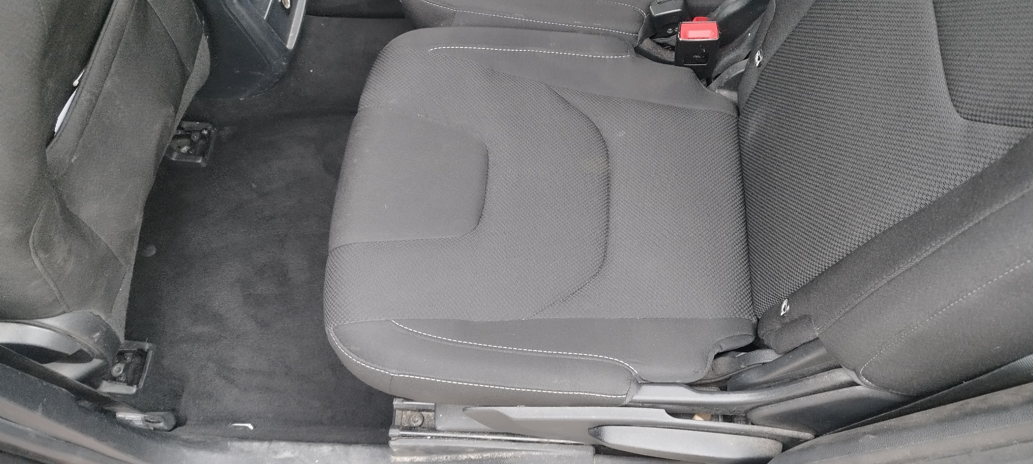 Szare tekstylne siedzenie samochodowe z białymi przeszyciami, widoczne mocowania do podłogi i czarna wykładzina.