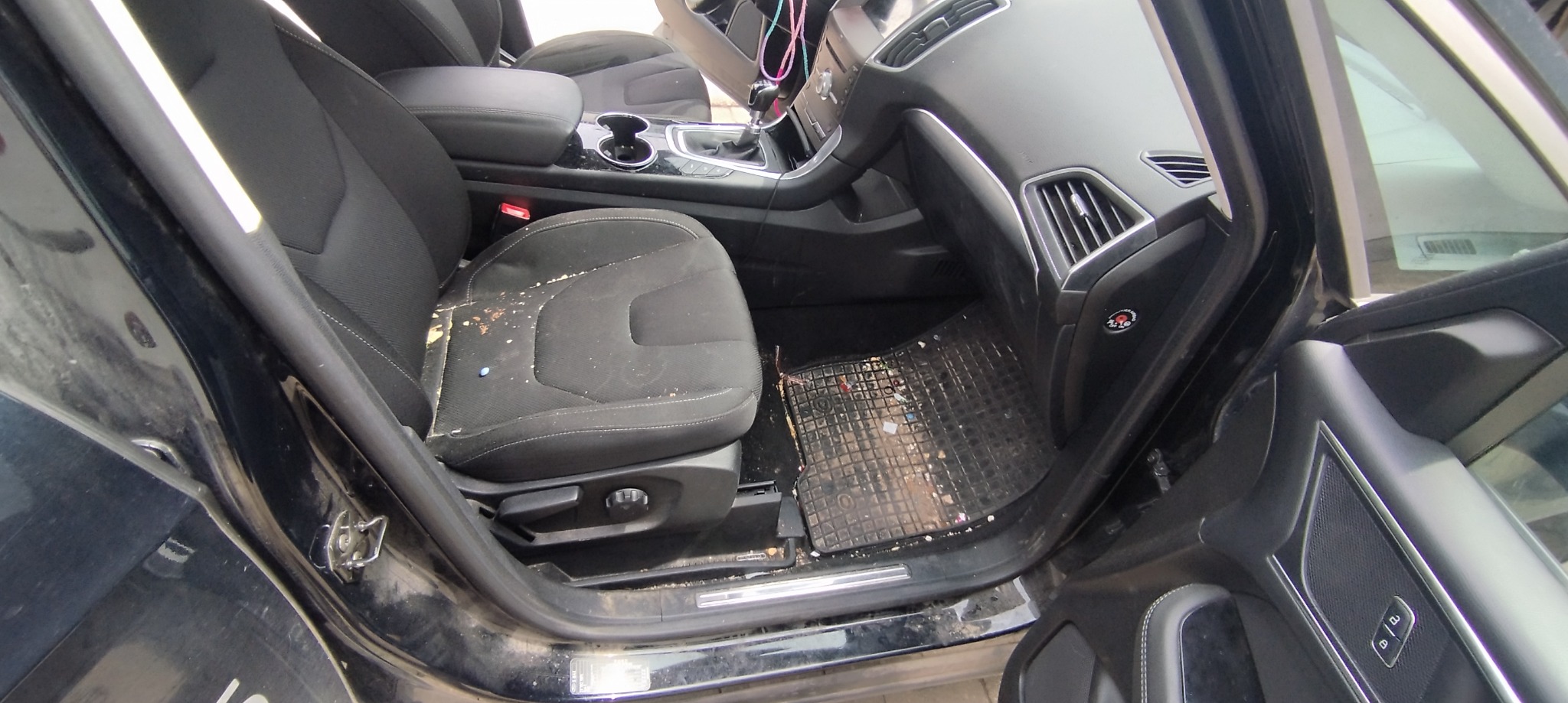 Wnętrze brudnego samochodu z widocznymi okruchami na siedzeniu, zabrudzonym dywanikiem i śladami użytkowania na desce rozdzielczej.