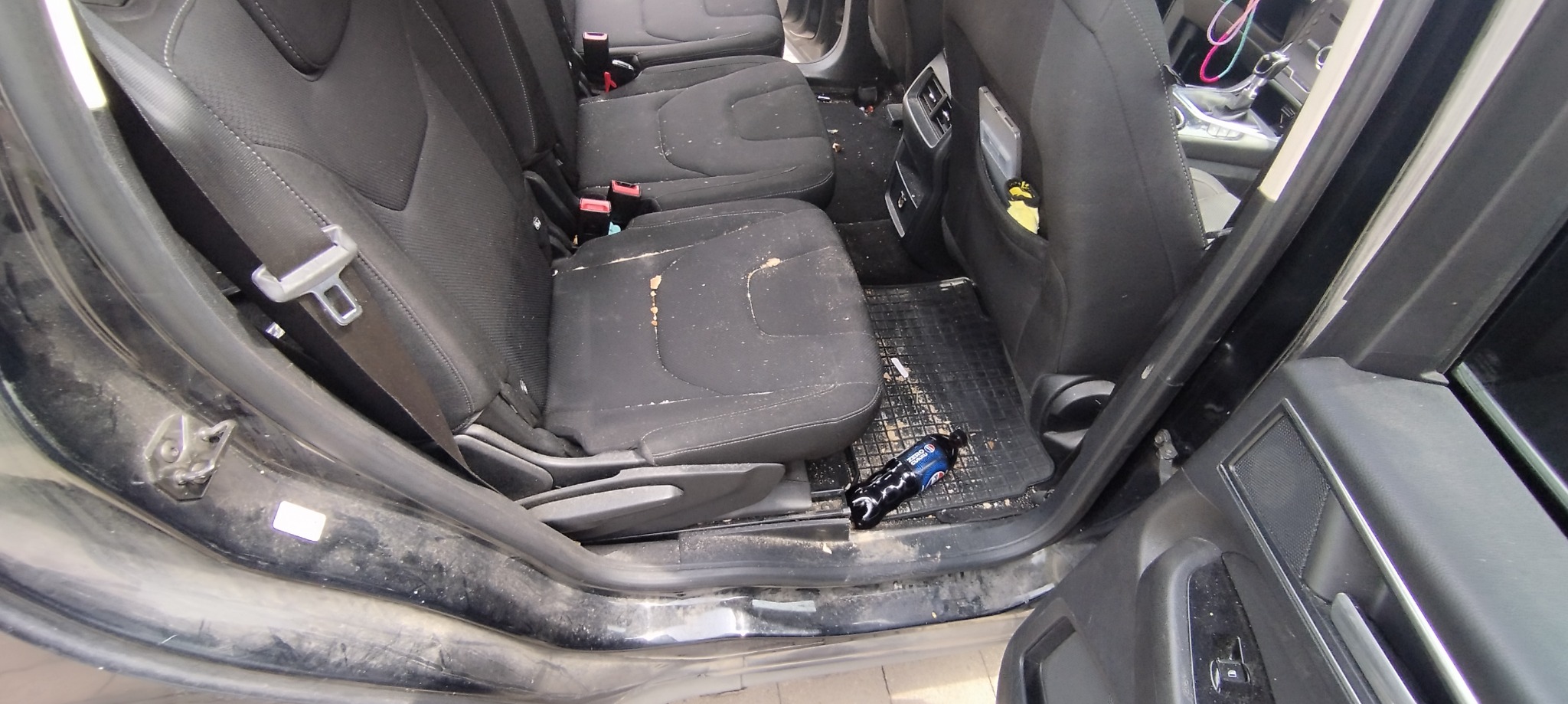 Wnętrze brudnego samochodu, widoczne tylne siedzenia pokryte okruchami i plamami, pusta butelka po napoju na podłodze przy progu.