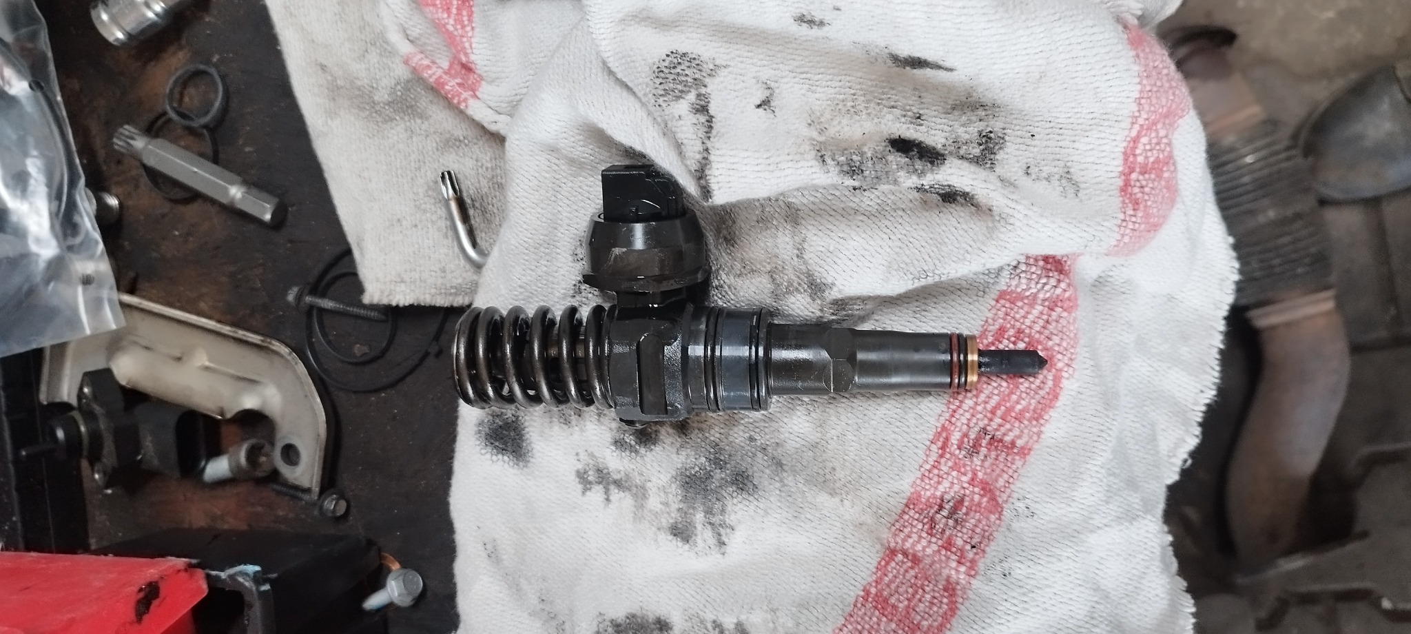 Brudny wtryskiwacz paliwa z widoczną sprężyną i końcówką, leżący na zabrudzonej szmatce warsztatowej, obok narzędzi.