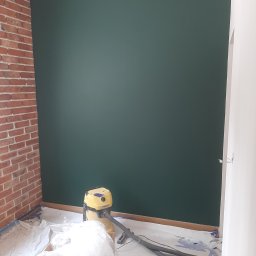 Mateusz Cichal Prace Wykończeniowe - Świeżo pomalowana, ciemnozielona ściana w pokoju z fragmentem ściany z czerwonej cegły, widoczny odkurzacz przemysłowy i folia malarska na podłodze, pozostałości po malowaniu.