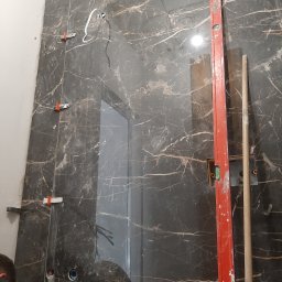 Mateusz Cichal Prace Wykończeniowe - Montaż płytek imitujących marmur na ścianie łazienki, widoczne poziomica, krzyżyki dystansowe, otwory na instalacje sanitarne.
