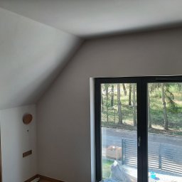 Mateusz Cichal Prace Wykończeniowe - Pokój na poddaszu po malowaniu ścian na jasny kolor, widoczne skośne ściany, okno z widokiem na drzewa i część regału.