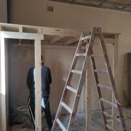 Mateusz Cichal Prace Wykończeniowe - Konstrukcja z drewnianych belek w trakcie montażu, widoczna drabina i pracownik w czarnej bluzie, przygotowanie do montażu płyt kartonowo-gipsowych.