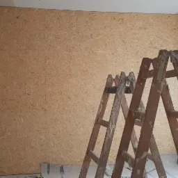 Ściana wyłożona płytą OSB w trakcie remontu, widoczna drewniana drabina, białe ściany wokół i fragment podłogi zabezpieczonej folią.