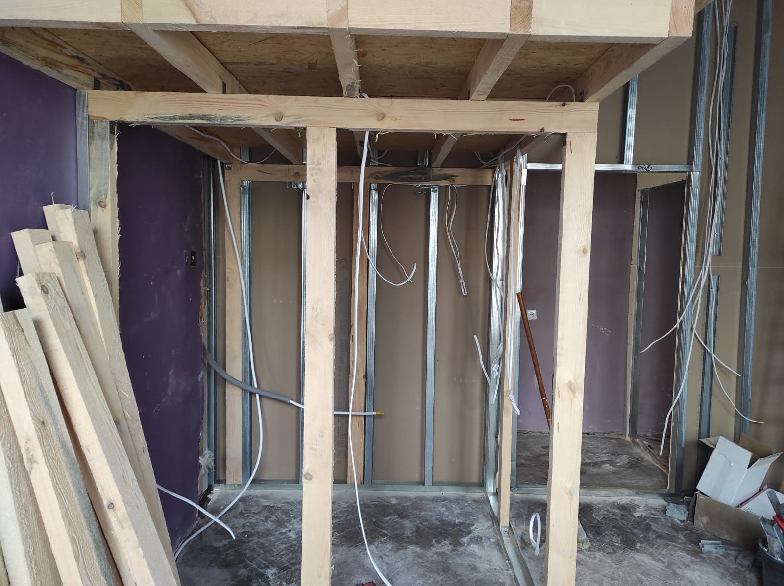 Wnętrze w trakcie prac: drewniana konstrukcja sufitu i ścianek działowych z profili stalowych, widoczne przewody elektryczne, stos desek oparty o fioletową ścianę.