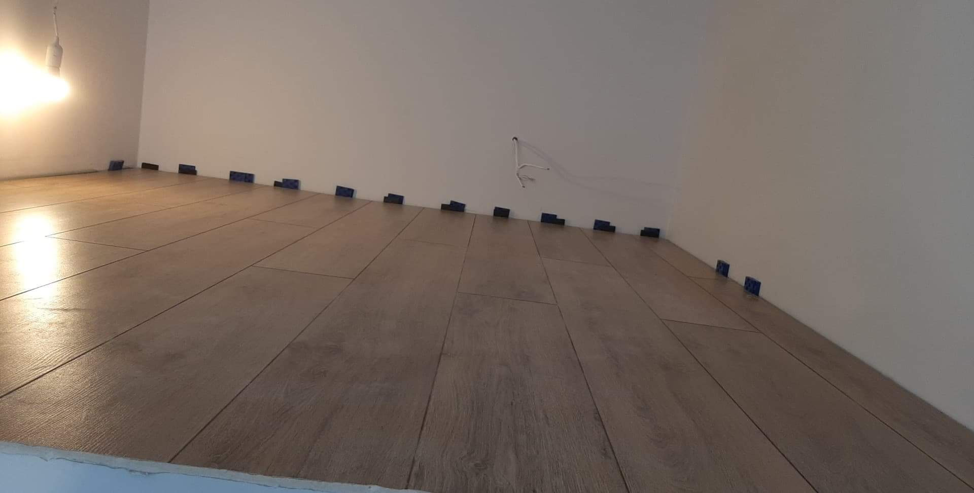 Układanie podłogi z paneli imitujących drewno, widoczne niebieskie kliny dystansowe wzdłuż ściany, surowa żarówka w tle.