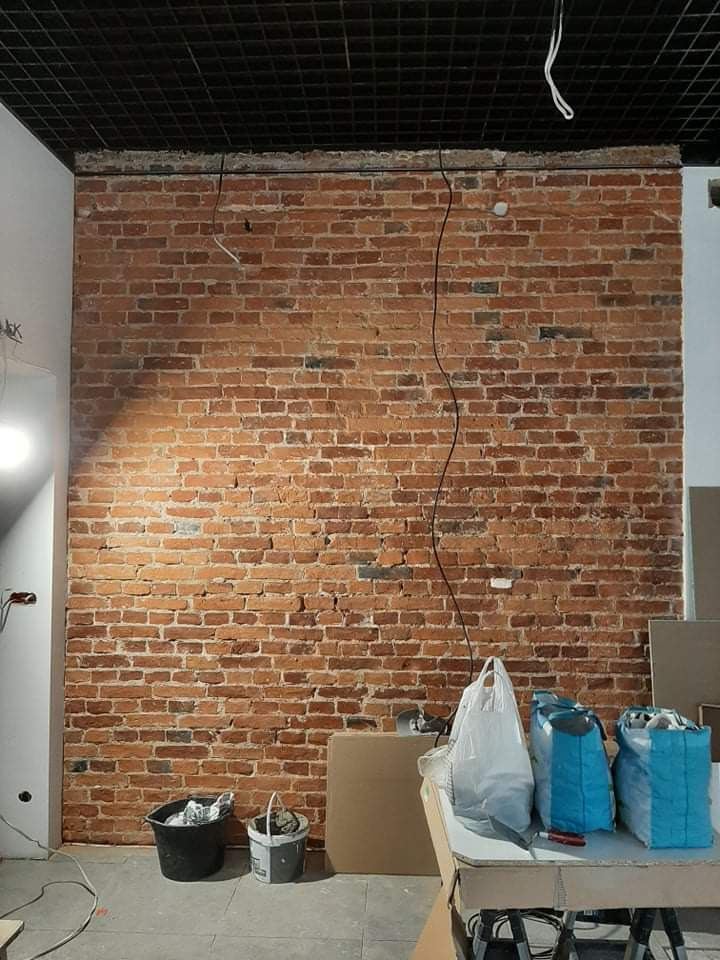 Ceglana ściana z widocznymi przewodami elektrycznymi, czarną kratownicą sufitową i elementami przygotowawczymi do prac wykończeniowych, takimi jak wiadra i worki z materiałami budowlanymi na stole.