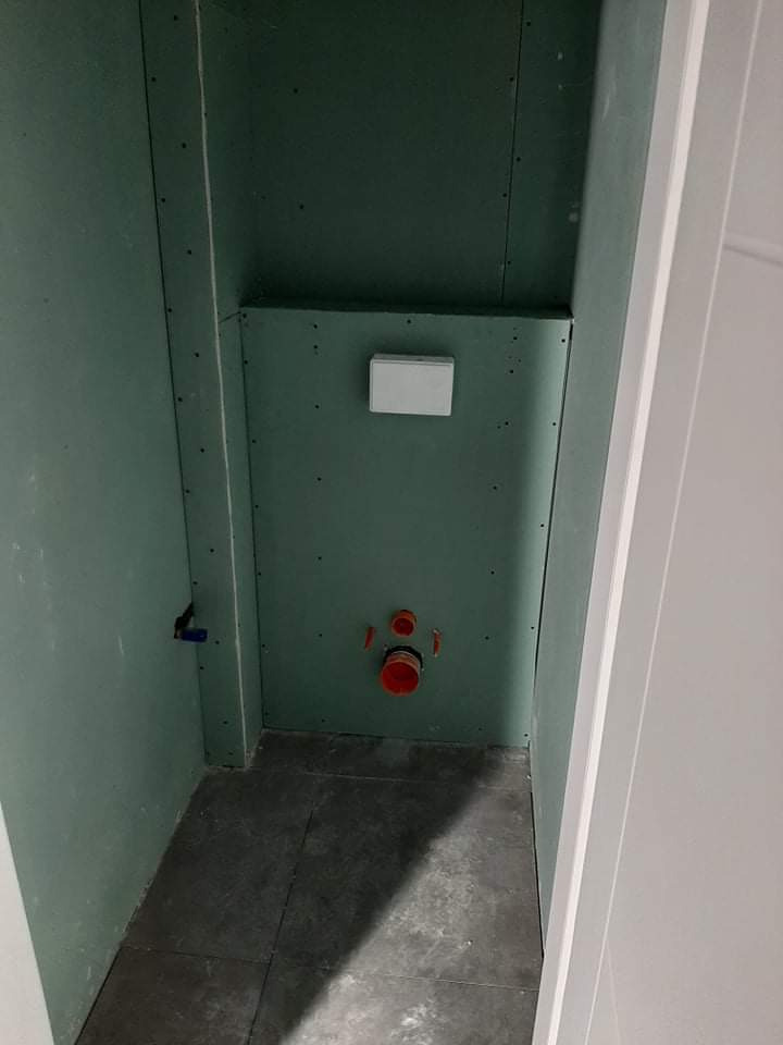 Montaż instalacji sanitarnej i zabudowa z płyty karton-gips w małej łazience, widoczne rury kanalizacyjne, szara podłoga z płytek i stelaż podtynkowy WC.