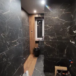 Wnętrze łazienki w trakcie remontu, ściany wyłożone czarnymi płytkami imitującymi marmur, widoczne narzędzia i materiały budowlane, fragment drewnianej podłogi i białe okno z żaluzjami.