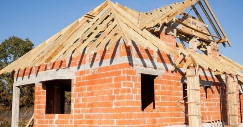 Surowy stan domu murowanego z czerwonej cegły ceramicznej, widoczne otwory okienne i zarys dachu z drewnianą konstrukcją krokwi, w tle drzewa i błękitne niebo.