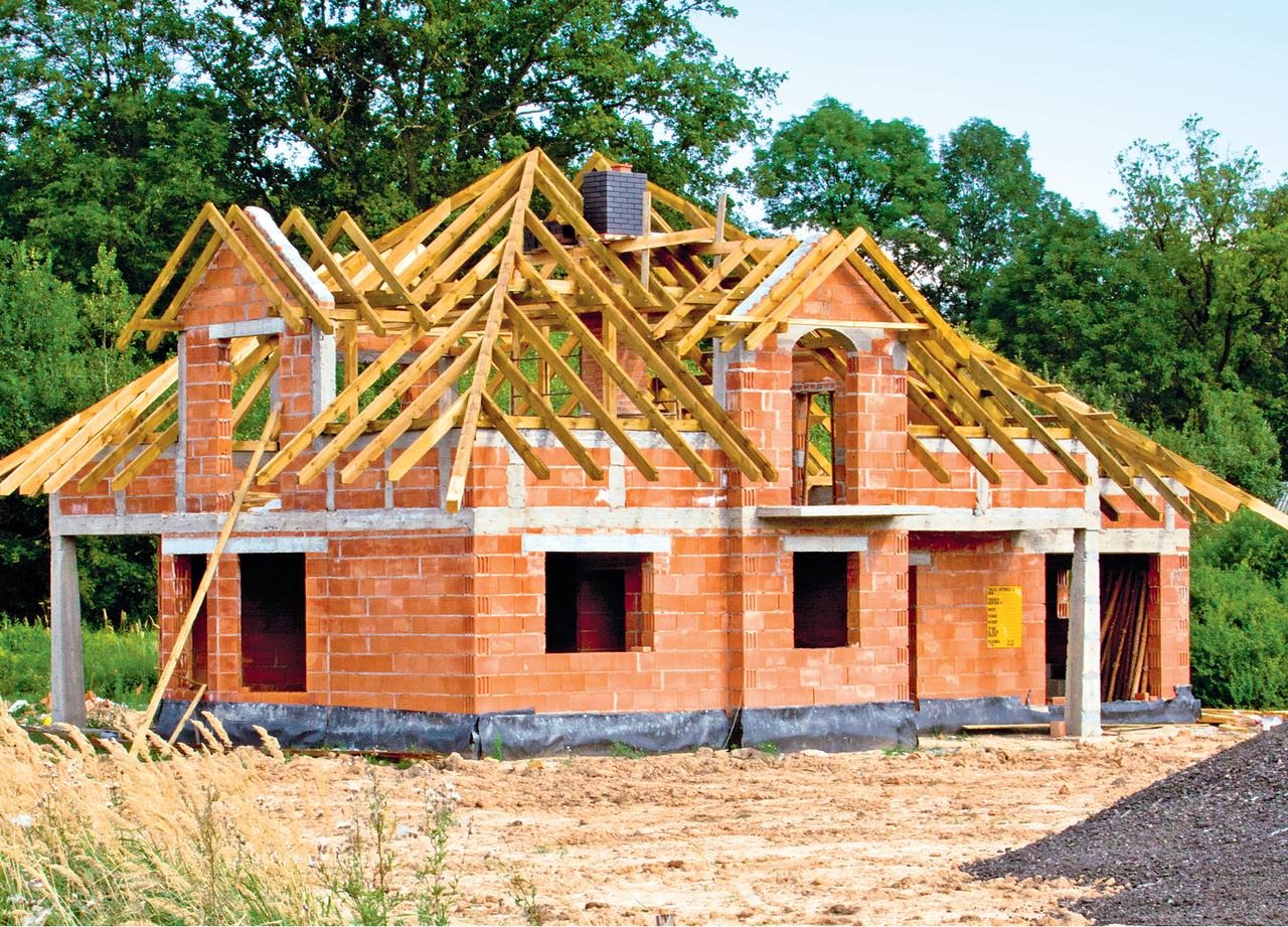 Surowy stan domu z czerwonej cegły z drewnianą konstrukcją dachu w trakcie budowy, widoczne otwarte okna i drzwi, w tle drzewa.