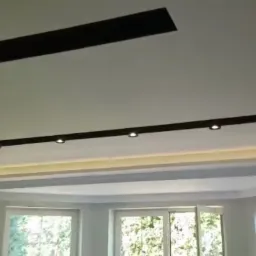 Biały sufit z wbudowanymi, czarnymi listwami oświetleniowymi i punktowymi lampami, widok na jasne pomieszczenie z oknami.