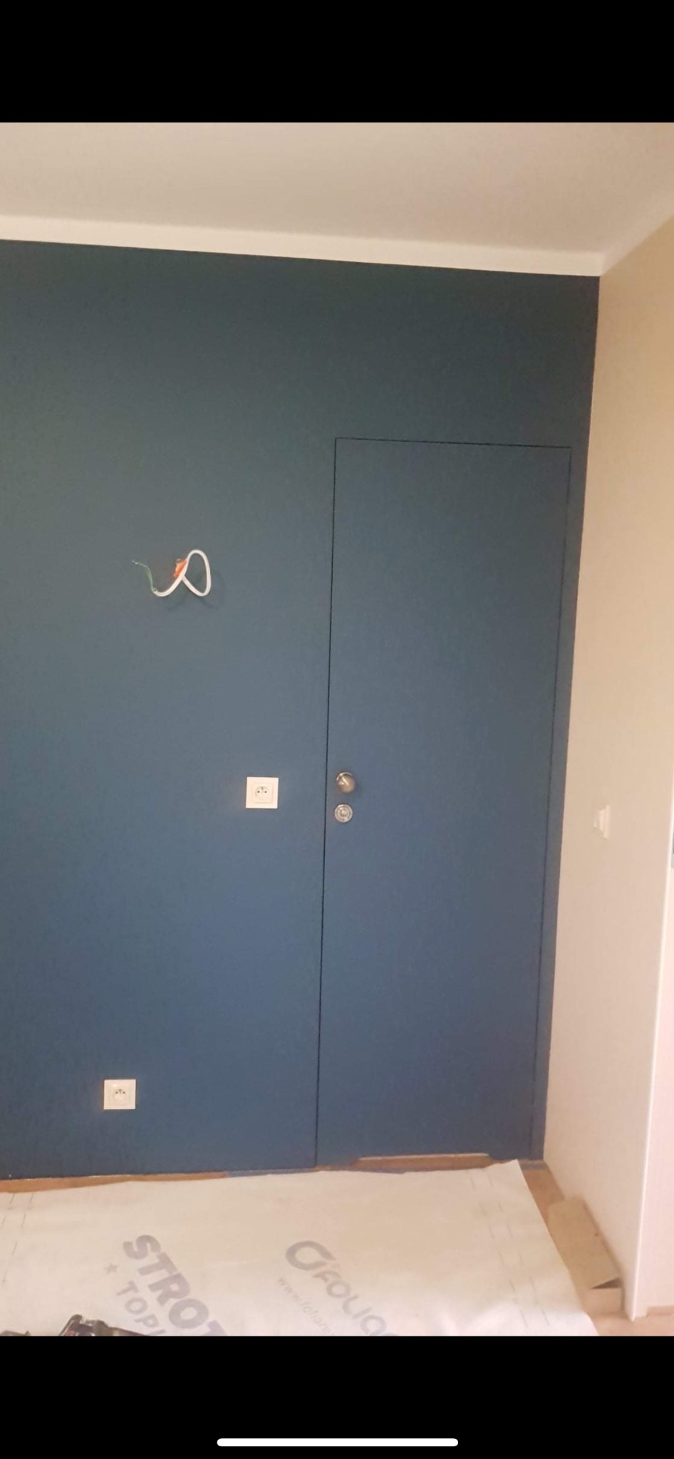 Niebieska ściana z ukrytymi drzwiami, widoczne gniazdka elektryczne i wystające kable, podłoga zabezpieczona folią ochronną.