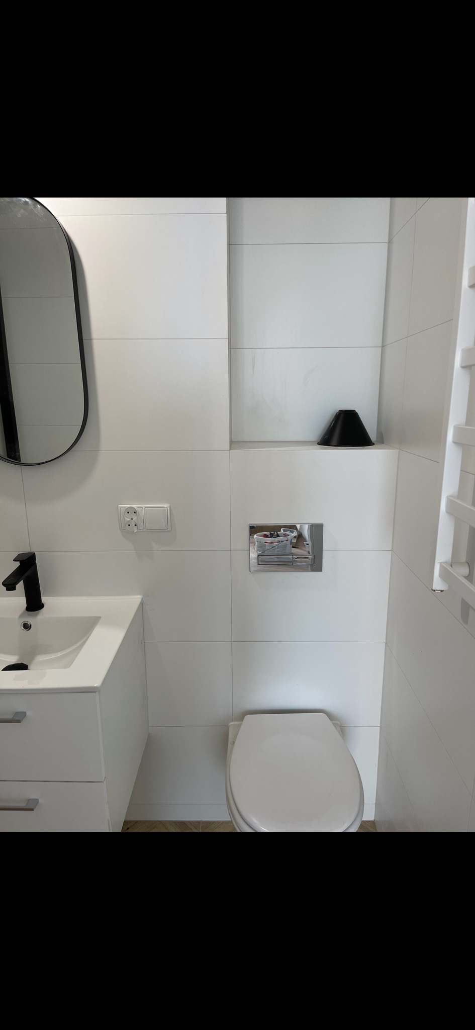 Minimalistyczna łazienka w bieli z czarnymi akcentami: umywalka z szafką, owalne lustro, wisząca toaleta, płytki ścienne i czarny stożek dekoracyjny we wnęce.