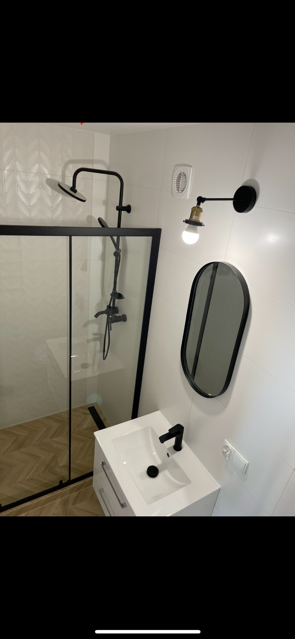 Nowoczesna łazienka z kabiną prysznicową typu walk-in z czarnymi ramami, białą umywalką z czarną armaturą, owalnym lustrem w czarnej ramie i designerską lampą ścienną z żarówką Edison