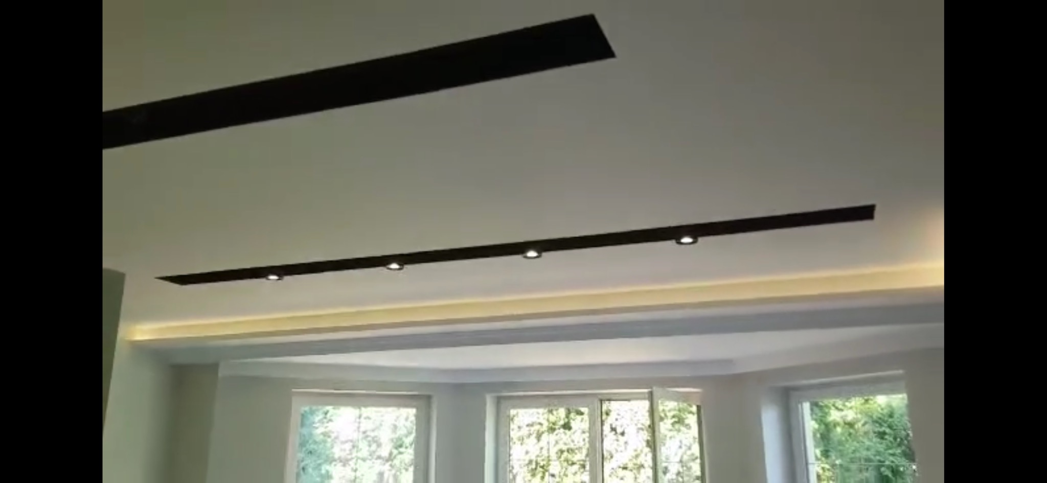 Biały sufit z wbudowanymi, czarnymi listwami oświetleniowymi i punktowymi lampami, widok na jasne pomieszczenie z oknami.