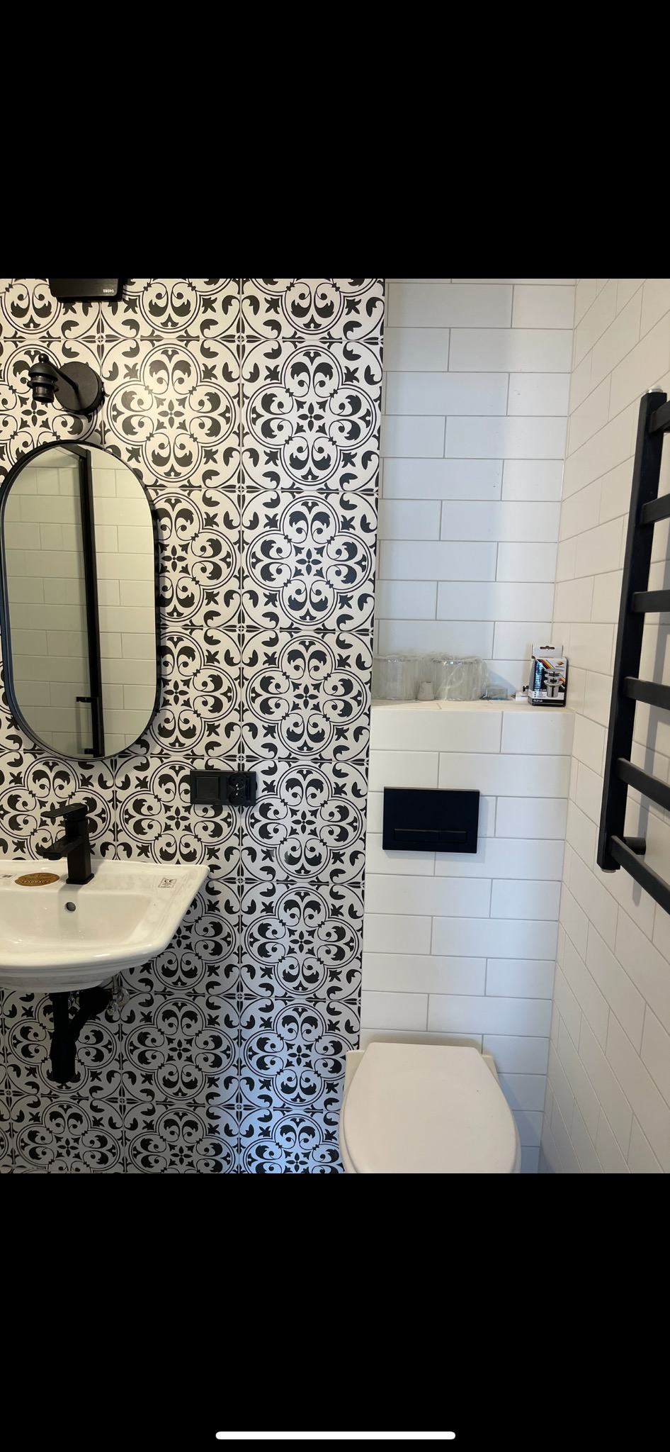 Nowoczesna łazienka z kontrastującym połączeniem płytek: wzorzyste czarno-białe płytki na jednej ścianie i białe prostokątne płytki na pozostałych ścianach, wisząca umywalka z czarną armaturą...