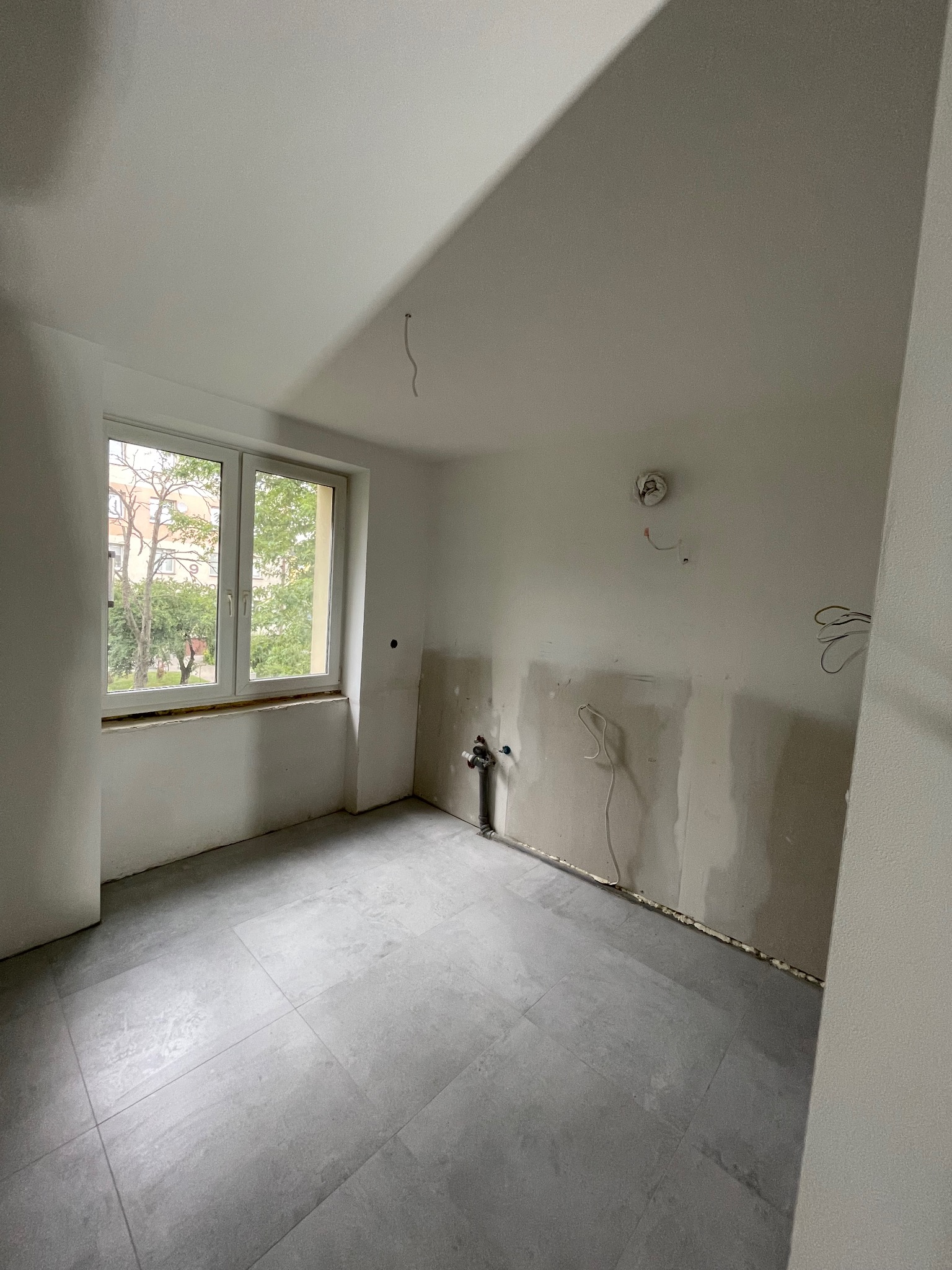 Wnętrze w trakcie remontu: szare płytki podłogowe, częściowo wykończone ściany z widocznymi przewodami elektrycznymi i rurą odpływową, okno z widokiem na zieleń.