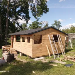 Surowa drewniana konstrukcja altany w trakcie budowy, z drabina opartą o ścianę i stertą odpadów drewnianych na trawie, w tle zieleń drzew i fragment murowanego budynku.