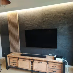 Ściana telewizyjna wykończona płytami imitującymi kamień z drewnianymi lamelami i podświetleniem LED, szafka RTV w stylu rustykalnym z przesuwnymi drzwiczkami.