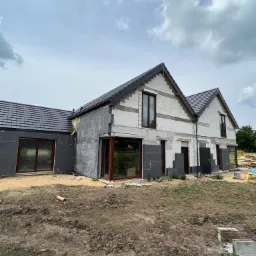 Dwukondygnacyjny dom jednorodzinny w trakcie ocieplania, widoczne fragmenty elewacji pokryte styropianem, okna z brązowymi ramami, ciemna dachówka, teren wokół domu częściowo przygotowany.