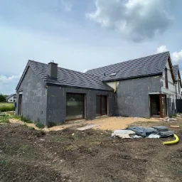 Dom jednorodzinny w trakcie ocieplania elewacji styropianem grafitowym, widoczne okna z brązowymi ramami, ciemna dachówka i przygotowany teren wokół budynku.