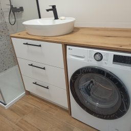 MEBLE TTR TOMASZ TRZEŚNIOWSKI - Nowoczesna łazienka z umywalką nablatową na blacie z drewnianym dekorem, szafką z trzema szufladami z czarnymi uchwytami oraz pralką LG w zabudowie.