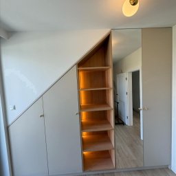 MEBLE TTR TOMASZ TRZEŚNIOWSKI - Szafa wnękowa z oświetlonymi półkami z drewna, dopasowana do skosu dachu, z lustrem wbudowanym w drzwi szafy, odbijającym wnętrze pokoju.