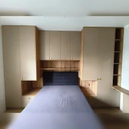 Drewniana zabudowa szafy wnękowej z półkami i szafkami nad łóżkiem z szarym zagłówkiem. Złote gałki. Realizacja na wymiar.