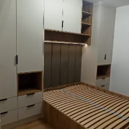 Nowoczesna sypialnia z zabudową meblową: białe szafy z czarnymi uchwytami, drewniany stelaż łóżka i tapicerowany zagłówek z oświetleniem LED.