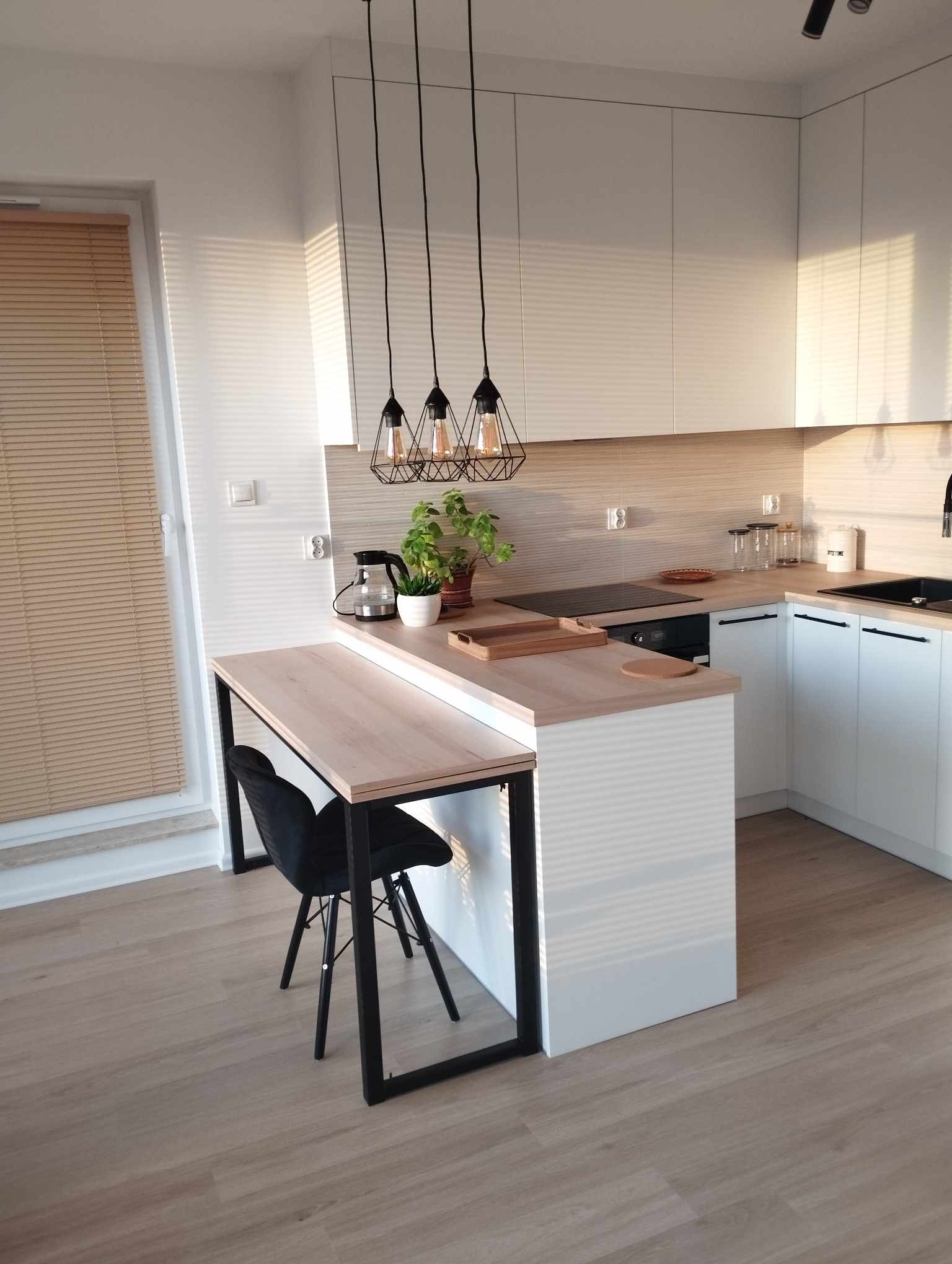 Jasna kuchnia z białymi szafkami, drewnianym blatem i wyspą z czarnym krzesłem, oświetlona trzema lampami wiszącymi w stylu industrialnym, z widokiem na okno z żaluzjami.