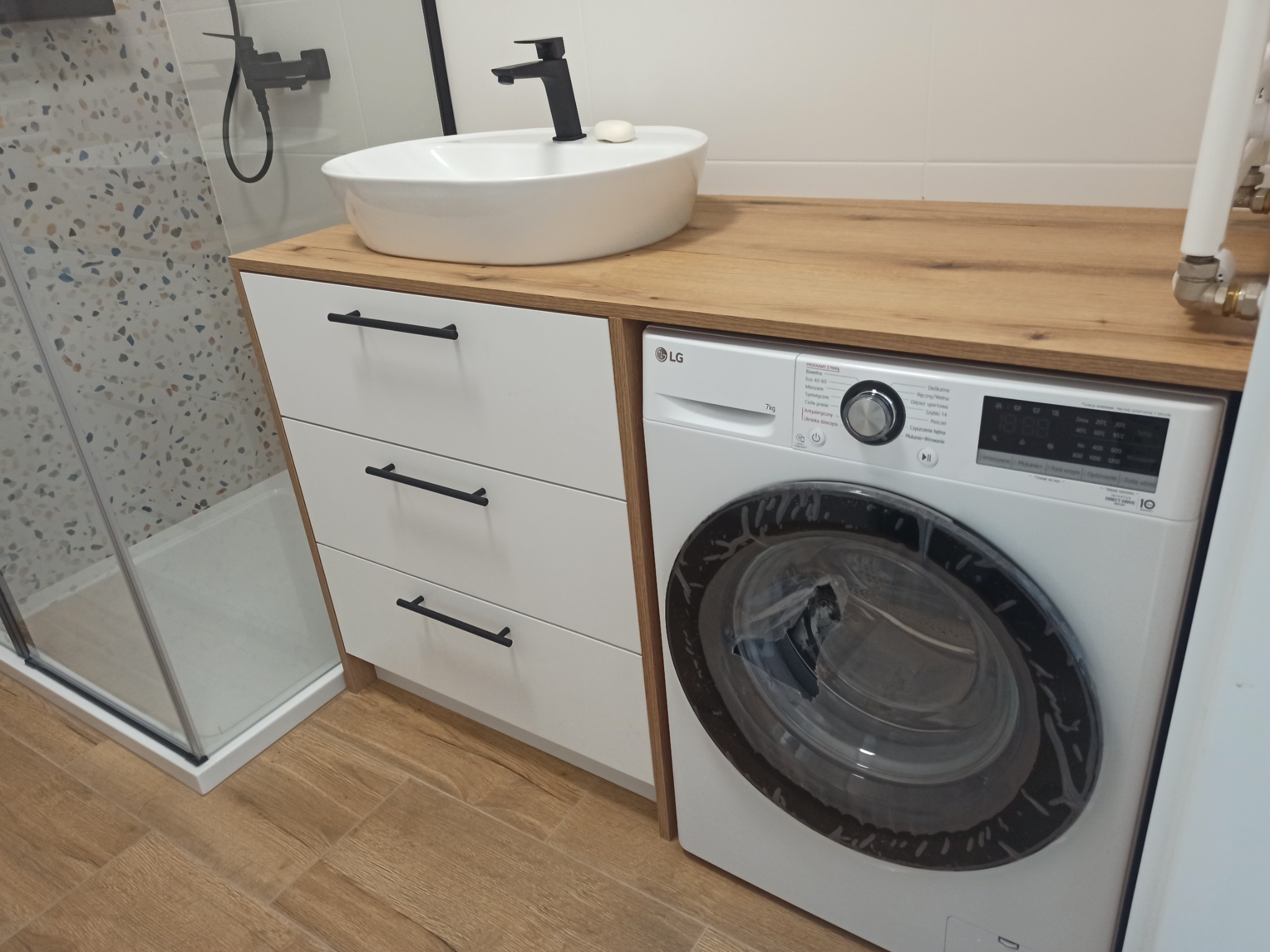 Nowoczesna łazienka z umywalką nablatową na blacie z drewnianym dekorem, szafką z trzema szufladami z czarnymi uchwytami oraz pralką LG w zabudowie.