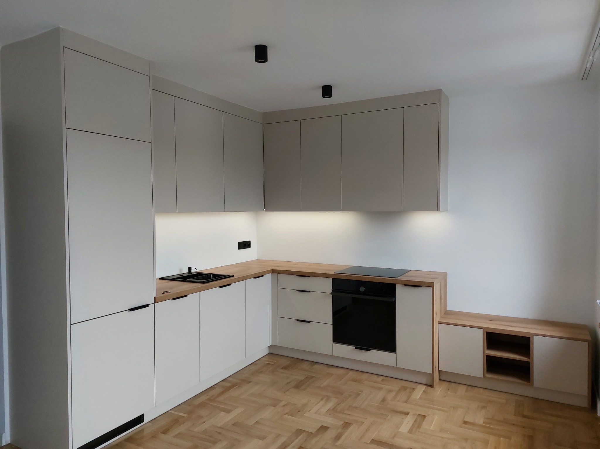 Jasna kuchnia na wymiar z drewnianym blatem i czarnymi akcentami, widok na parkiet w jodełkę.