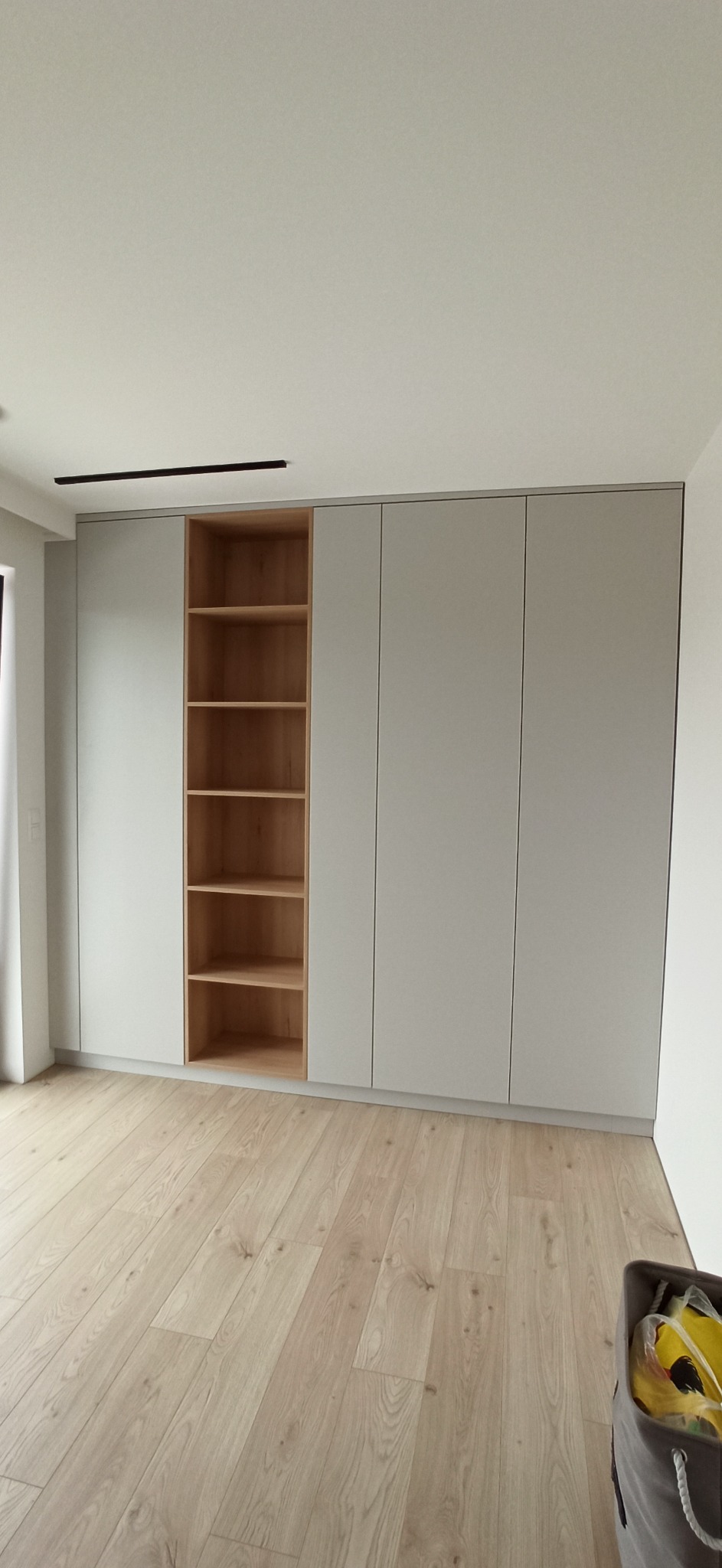 Szara szafa wnękowa z drewnianymi półkami, widok na jasną podłogę i fragment kosza na pranie.