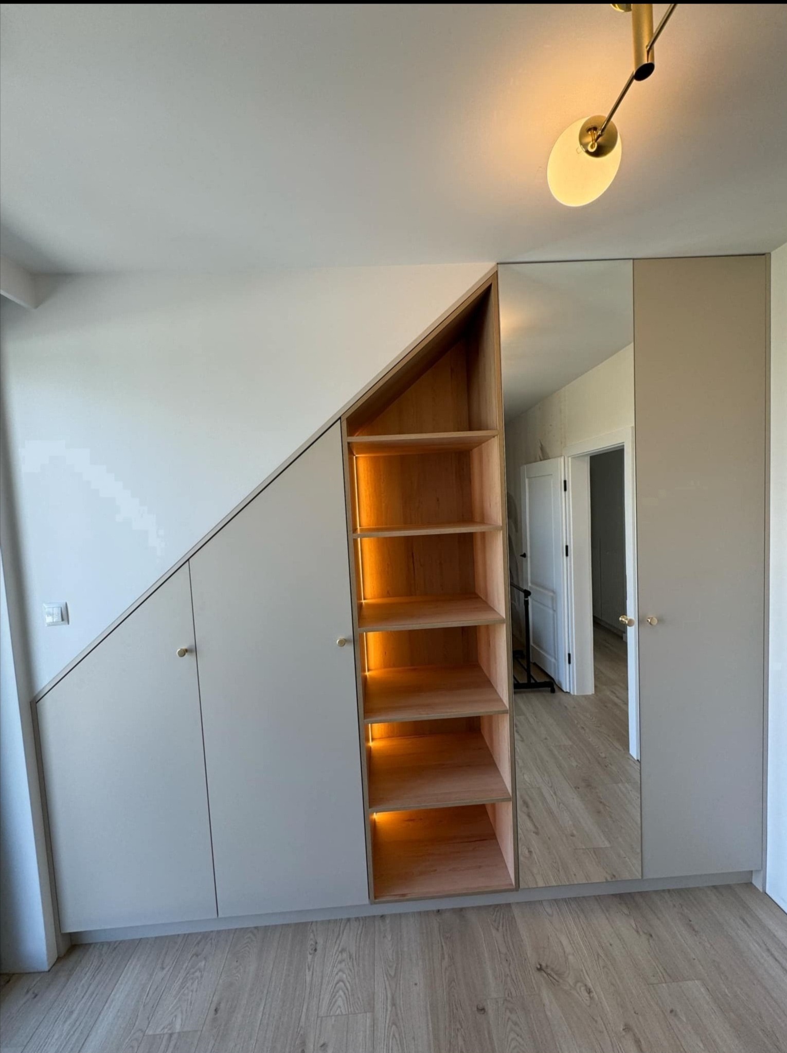 Szafa wnękowa z oświetlonymi półkami z drewna, dopasowana do skosu dachu, z lustrem wbudowanym w drzwi szafy, odbijającym wnętrze pokoju.