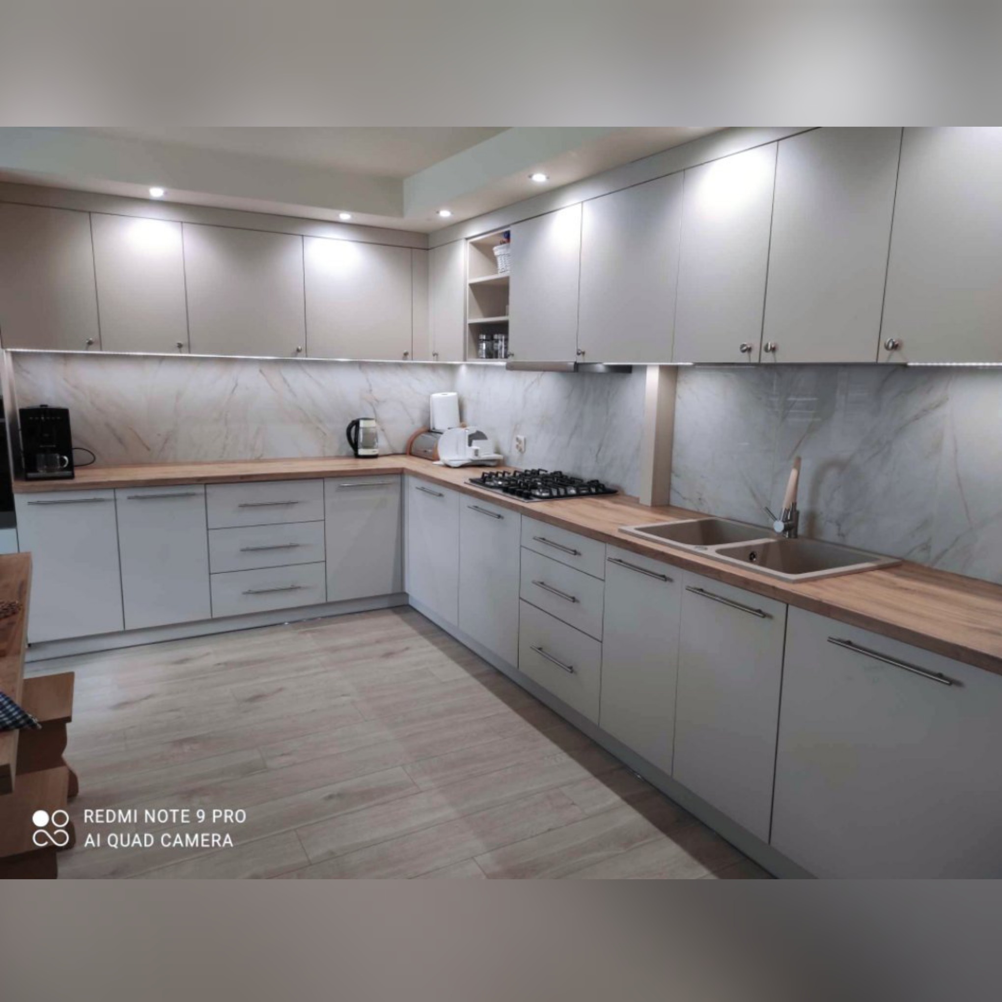 Jasnoszara kuchnia z drewnianym blatem, marmurowym backsplashiem i oświetleniem LED pod szafkami wiszącymi.