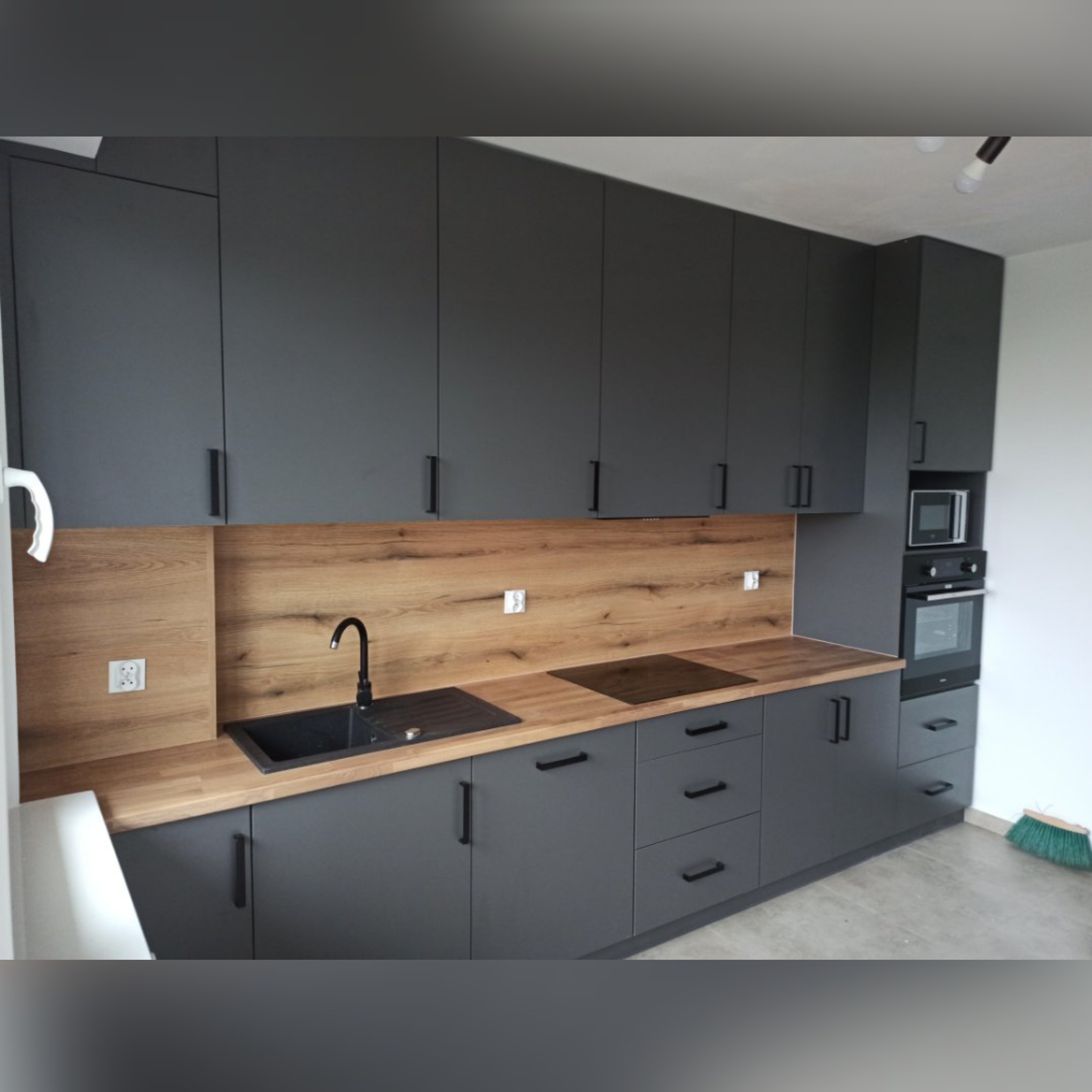 Nowoczesna kuchnia w odcieniach szarości z drewnianym blatem i panelem ściennym, czarny zlew i płyta indukcyjna, minimalistyczne uchwyty, oświetlenie punktowe, widoczna część ściany i miotła.