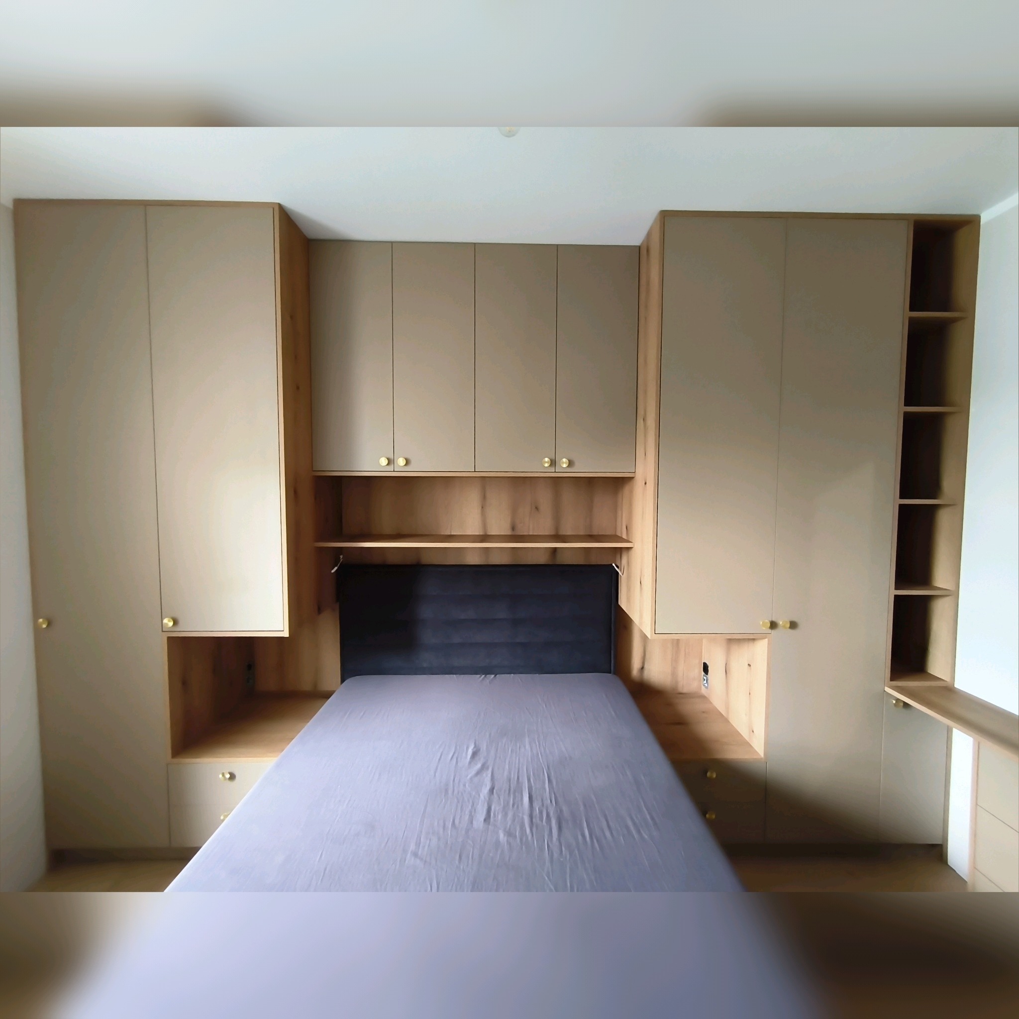 Drewniana zabudowa szafy wnękowej z półkami i szafkami nad łóżkiem z szarym zagłówkiem. Złote gałki. Realizacja na wymiar.