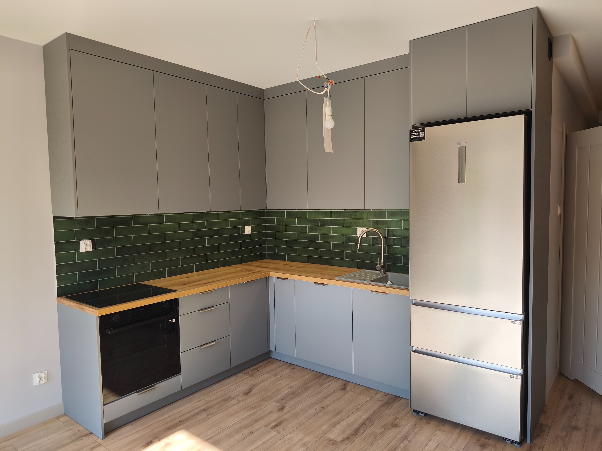 Nowoczesna kuchnia w odcieniach szarości z drewnianym blatem i zielonymi płytkami. Widoczna zabudowa kuchenna, lodówka i płyta grzewcza. Surowa żarówka zwisa z sufitu.