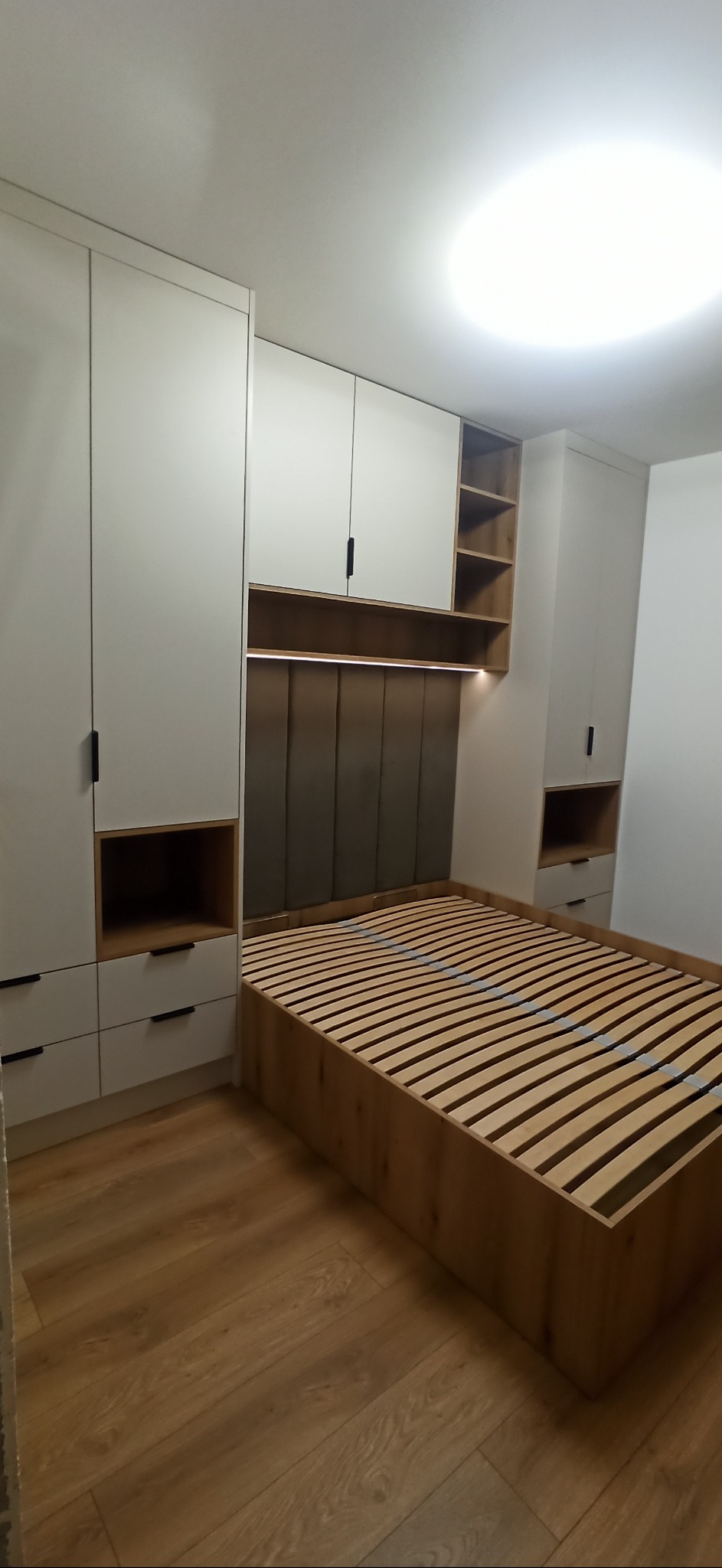 Nowoczesna sypialnia z zabudową meblową: białe szafy z czarnymi uchwytami, drewniany stelaż łóżka i tapicerowany zagłówek z oświetleniem LED.