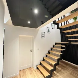 Błyszczący, czarny sufit napinany odbija schody z drewnianymi stopniami i czarną konstrukcją w nowoczesnym holu z białymi ścianami i drewnianą podłogą.