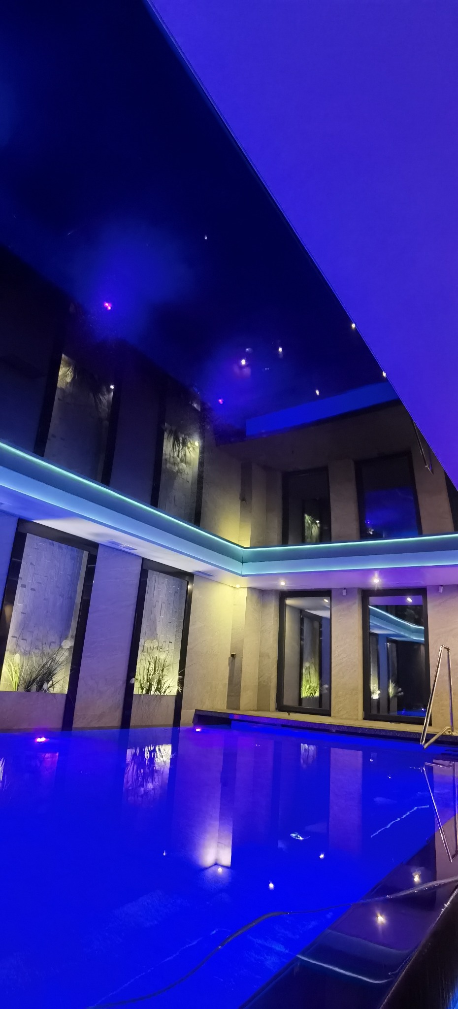 Błękitny basen z ciemnym, błyszczącym sufitem z punktowym oświetleniem, odbijającym się w wodzie, otoczony ścianami z wnękami z roślinami i podświetleniem LED.